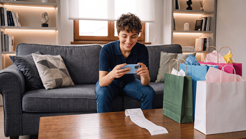 Una giovane donna sorridente seduta sul divano di un soggiorno moderno mentre usa lo smartphone per fotografare uno scontrino d'acquisto appoggiato sul tavolo, accanto a diversi sacchetti regalo colorati.